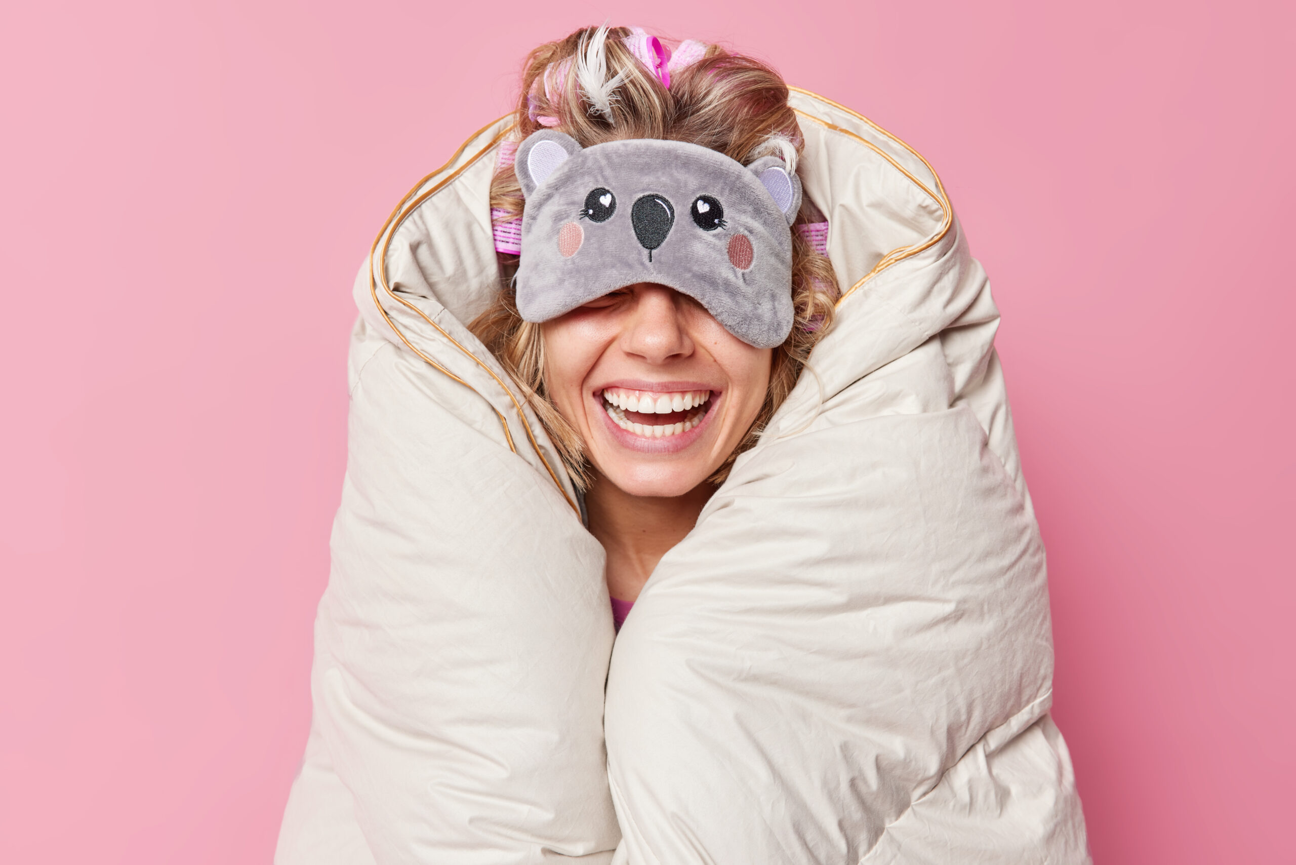 Unrecognizable cheerful woman wears sleepmask on eyes smiles broadly wrapped in duvet expresses positive emotions being in good mood isolated over pink background. People rest and bedtime concept