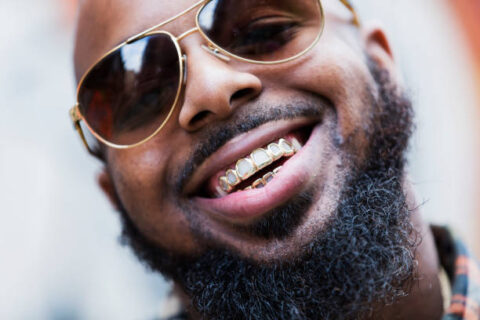 An African American man with a gold grill, thick beard and sunglasses, smiling.